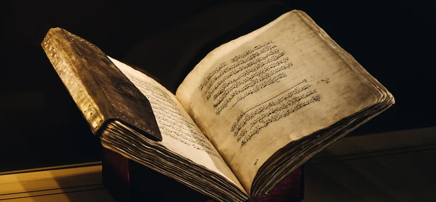 An old book in a leather binding lies open on a stand, its yellowed pages handwritten in black Arabic script, lit against a dark background