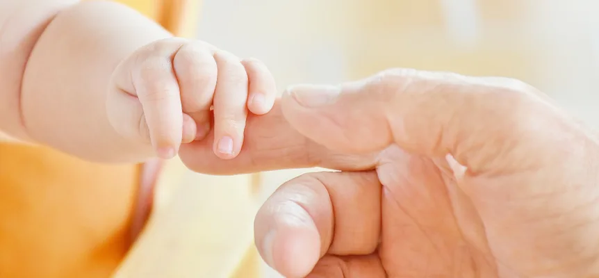 A baby's hand wrapped around an adult's finger against a pale yellow background