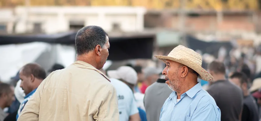 Deux hommes qui discutent dans un marché en plein air bondé, l'un en veste beige, à moitié de dos, l'autre en chapeau de paille et chemise bleu clair