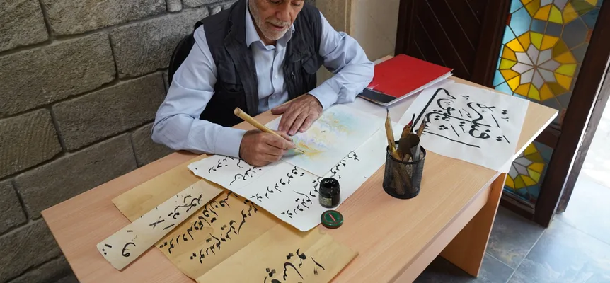 An older man writing Arabic calligraphy with a reed pen at a wooden desk covered in practice sheets, beside a stained-glass window