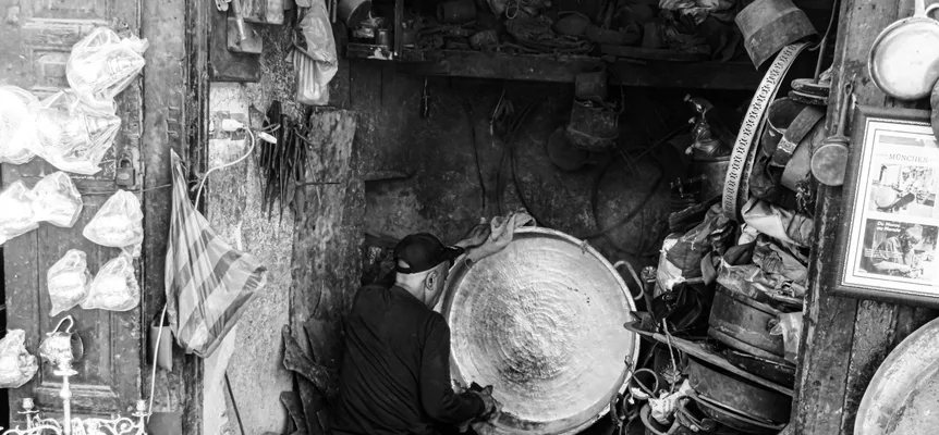 Black-and-white photo of a metalworker in a cap polishing a large hammered metal basin, with pots and pans piled around his workshop in Fez