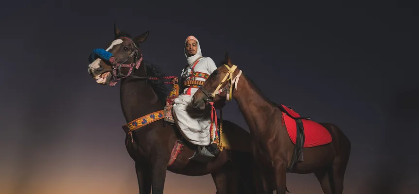 A man in a white robe and headscarf on a dark horse with an embroidered saddle and bridle, a second saddled horse beside him, against a dusk sky