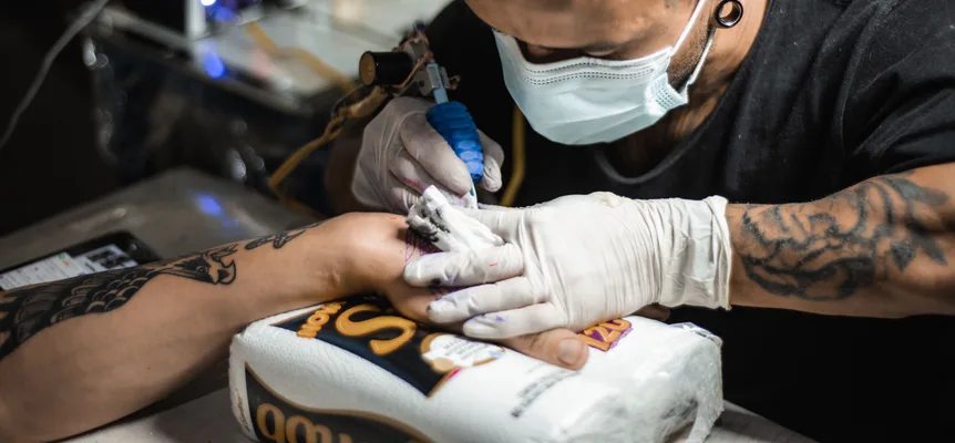 A tattoo artist in a face mask and white gloves inks a design on a client's hand, which rests on a roll of paper towels