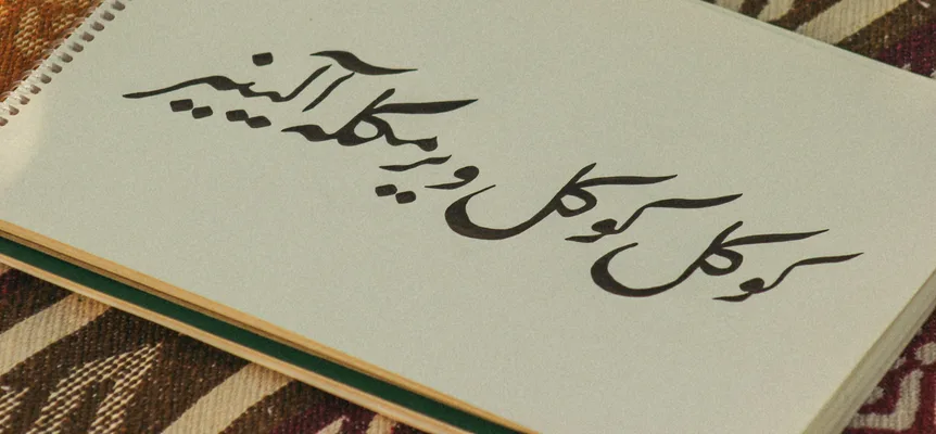 A spiral sketchbook lying on a woven rug, its page filled with black ink calligraphy in the Arabic script