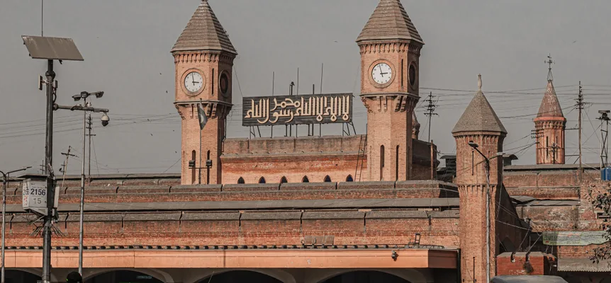 The red-brick front of a railway station in Lahore, with two clock towers, a sign in Arabic calligraphy between them, and traffic and crowds in the street below