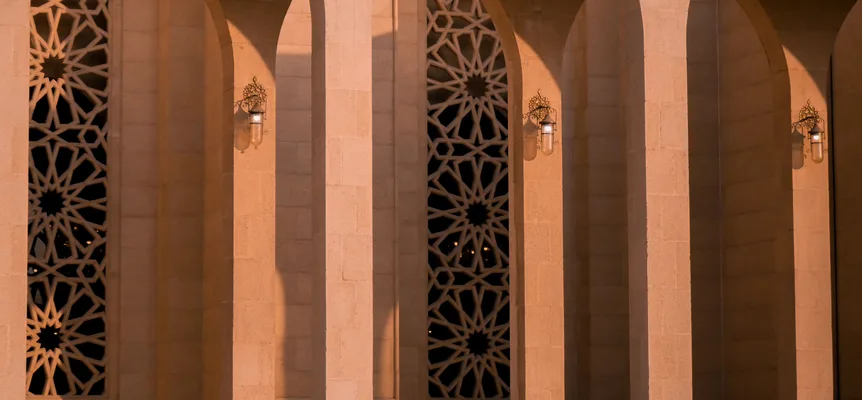 A row of sandstone arches in warm evening light, with dark star-pattern lattice screens between them and a brass lamp on each pillar