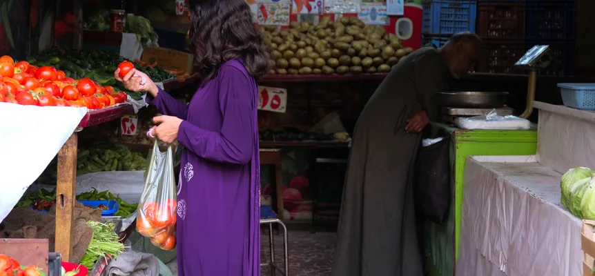 A woman in a purple dress picks tomatoes into a plastic bag at a Cairo vegetable stall while an older man in a grey galabiya weighs produce at the back
