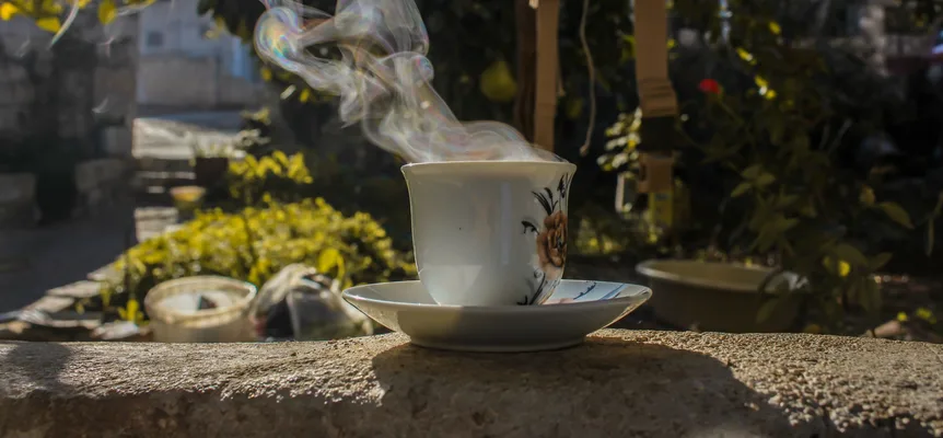 Steam rising from a white cup painted with a rose, standing on a saucer on a stone wall in a sunny garden