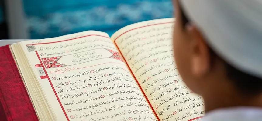 Seen over the shoulder of a boy in a white cap, an open Quran with red-bordered pages and Arabic text