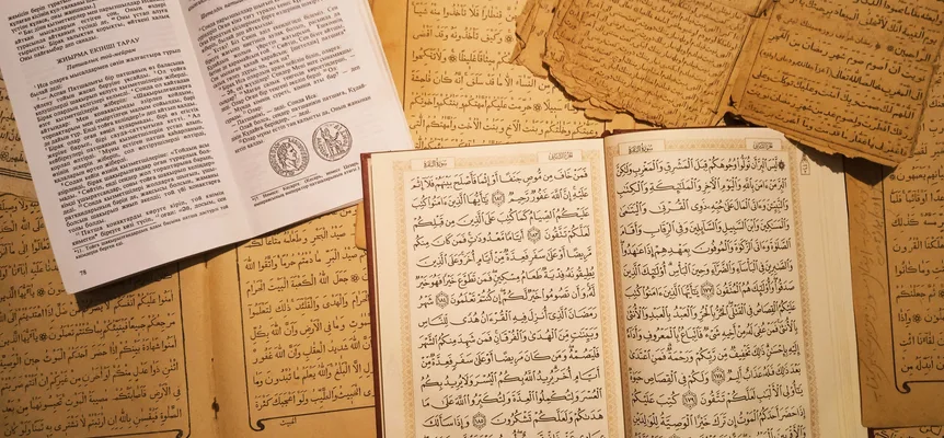 An open printed Quran lying on loose, yellowed handwritten pages in Arabic script, beside an open book printed in Cyrillic letters