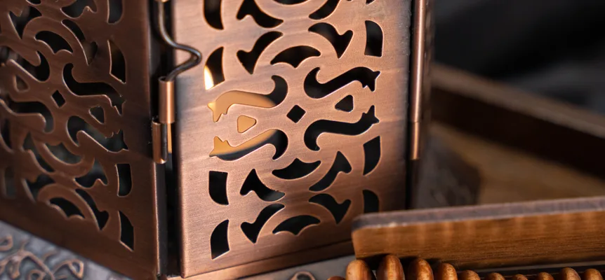 Candlelight through the cut-out panels of a copper lantern, beside a string of wooden prayer beads